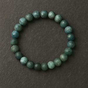 May include: A teal-colored beaded bracelet. The bracelet is composed of round, matte beads in varying shades of teal and green. The beads are strung together to form a circular shape, resting on a dark gray surface.