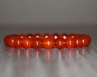May include: A vibrant orange beaded bracelet. The beads are round and appear to be made of a translucent material, reflecting light. The bracelet is displayed on a light-colored surface, with a soft, blurred background.