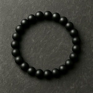 May include: A matte black beaded bracelet. The bracelet is composed of numerous round, dark beads, arranged in a circular pattern. The beads have a smooth, non-reflective surface, creating a uniform, dark appearance. The bracelet is displayed on a dark gray surface.