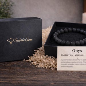 May include: A black onyx bead bracelet displayed in an open black box with a gold logo. A card with the word "Onyx" and the words "Protection, Strength, Stability" is next to the bracelet. The box is on a wooden surface.