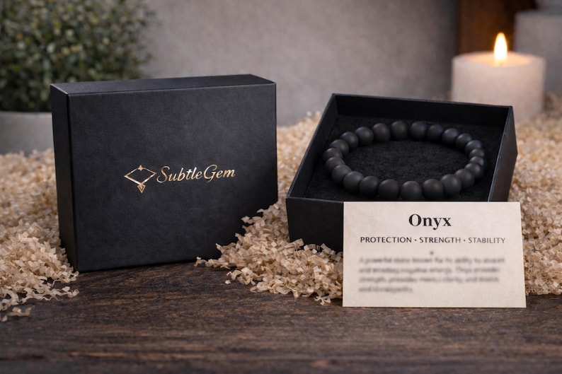 May include: A black onyx bead bracelet displayed in an open black box with a gold logo. A card with the word "Onyx" and the words "Protection, Strength, Stability" is next to the bracelet. The box is on a wooden surface.
