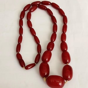 Puede incluir: Un collar vintage con grandes cuentas ovaladas de color rojo intenso. Las cuentas están ensartadas con un cordón de color neutro, creando un accesorio llamativo. El collar es una pieza llamativa.
