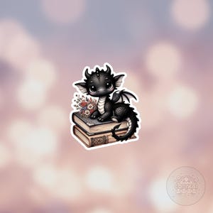 Puede incluir: Un pequeño dragón negro con cuernos y alas blancas se sienta encima de dos libros antiguos apilados. El dragón tiene un ramo de flores detrás.