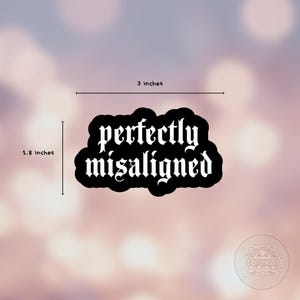 May include: A black sticker with the words "perfectly misaligned" in a gothic font. The sticker is 3 inches wide and 1.8 inches tall. The background is a blurred mix of pink and white.
