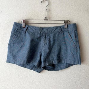 May include: Blue denim shorts featuring a mid-rise waist, a button and zip fly closure, two front slash pockets, and belt loops. The fabric has a subtle heathered texture with a slightly faded appearance, hanging from a metal hanger.