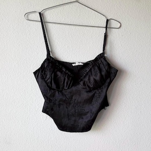 May include: Black satin bustier top with adjustable spaghetti straps. The top features a floral pattern and a sweetheart neckline. The garment is hanging on a metal hanger against a white wall.