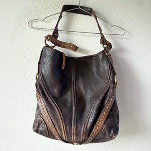 May include: A dark brown leather hobo bag featuring a textured surface, vertical stitched panels, and decorative brass-toned metal studs along the front straps. The bag includes a single shoulder strap attached with large brass-toned metal rings.