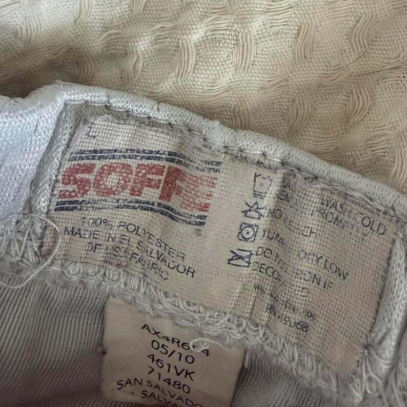 May include: A white clothing tag with the red Soffe logo, stating 100% polyester and made in El Salvador. The tag includes care instructions such as wash cold, do not bleach, tumble dry low, and do not iron. A secondary tag shows codes AX4R6-4, 05/10, 461VK, 71480.