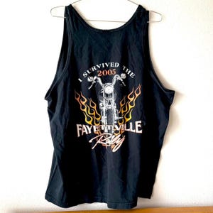May include: Black tank top with a motorcycle graphic and the text "I Survived the 2005 Fayetteville Rally". The graphic features a motorcycle with flames. The tank top is hanging on a metal hanger.