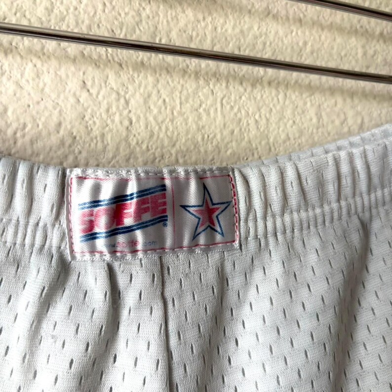 May include: White athletic shorts made of a breathable mesh fabric with an elastic waistband. A rectangular white fabric label is sewn onto the center front, featuring the red text SOFFE and a blue star outline with a red center star graphic.