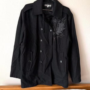 May include: Black double-breasted coat with a decorative emblem on the chest. The coat features a notched collar, shoulder epaulets, and two front pockets. The coat has six buttons and a brand label at the neck.