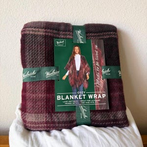 Puede incluir: Una manta envolvente Woolrich plegada con un patrón de cuadros en color burdeos oscuro y marrón. Una etiqueta verde está fijada en el centro, mostrando el texto Woolrich, Cozy Blanket Wrap, Luxe Softness, Fringe Detail y One Size Fits All.