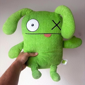 May include: A bright green plush toy with a rectangular body, two long floppy ears, and two short arms and legs. The face features one large white circular eye with a black pupil, one black stitched X for an eye, and a small pink tongue.