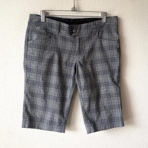 May include: Grey and black plaid shorts featuring a mid-length cut, a front button closure with two dark buttons, two side pockets, and a black interior waistband lining. The garment is displayed on a metal hanger against a plain white wall.