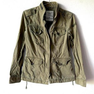 May include: An olive green jacket with multiple pockets and a zip-up front. The jacket features a stand-up collar and a drawstring at the waist. The brand name "Abercrombie & Fitch" is visible on a tag inside. A stylish, casual outerwear piece.