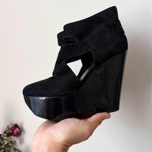 May include: Black wedge platform shoe with a suede upper and a glossy platform sole. The shoe features a cut-out design on the side and a strap closure. The heel is approximately 5 inches high.