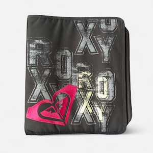 Puede incluir: Un archivador negro con la palabra "ROXY" en una fuente desgastada y texturizada. El logotipo de Roxy, un diseño rosa en forma de corazón, está en la parte inferior izquierda. El archivador tiene un cierre de cremallera.