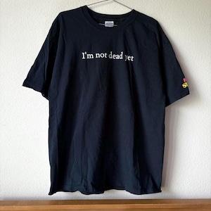 May include: A black short-sleeved t-shirt featuring the white text "I'm not dead yet" printed across the chest. The shirt has a crew neckline and a small graphic detail in red and yellow on the left sleeve, hanging on a metal hanger.