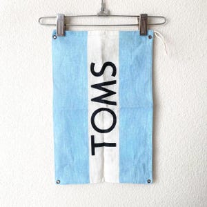 Pode incluir: Bandeira retangular azul e branca com a palavra "TOMS" impressa verticalmente em preto. A bandeira está pendurada em um cabide de metal. A bandeira tem um fundo azul claro com uma faixa branca vertical no centro.