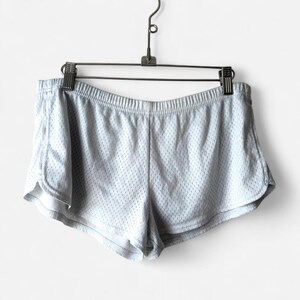 May include: Light gray athletic shorts featuring a breathable mesh fabric construction, an elasticized waistband, and curved hemline detailing. The shorts are suspended from a metal hanger with two clips, showcasing the lightweight material.