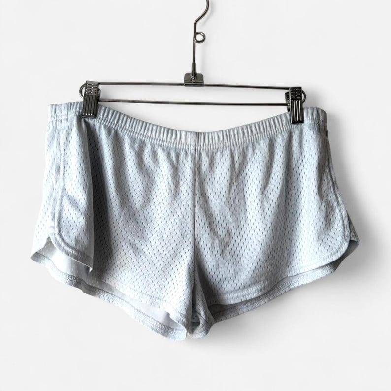 May include: Light gray athletic shorts featuring a breathable mesh fabric construction, an elasticized waistband, and curved hemline detailing. The shorts are suspended from a metal hanger with two clips, showcasing the lightweight material.