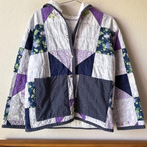 May include: A quilted hooded jacket featuring a patchwork design of purple, navy blue, and white floral fabrics. The garment includes two large front pockets made of navy blue fabric with white polka dots and a front metal snap button closure.