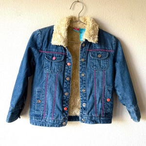 May include: A blue denim jacket featuring a cream-colored faux fur collar and interior lining. The jacket includes two chest pockets with flaps, vertical pink decorative stitching, and a front button closure with alternating silver and orange buttons.