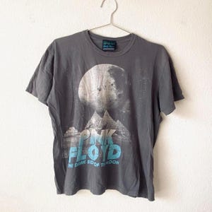 May include: Gray t-shirt featuring a large moon graphic above a mountain range and the words "PINK FLOYD" in blue. Below the band name is the text "THE DARK SIDE OF THE MOON". The shirt is hanging on a metal hanger.