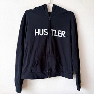 May include: Black zip-up hoodie with the word "HUSTLER" printed in white across the chest. The hoodie features a hood, a front pocket, and a full-length zip. The garment is hanging on a metal hanger.