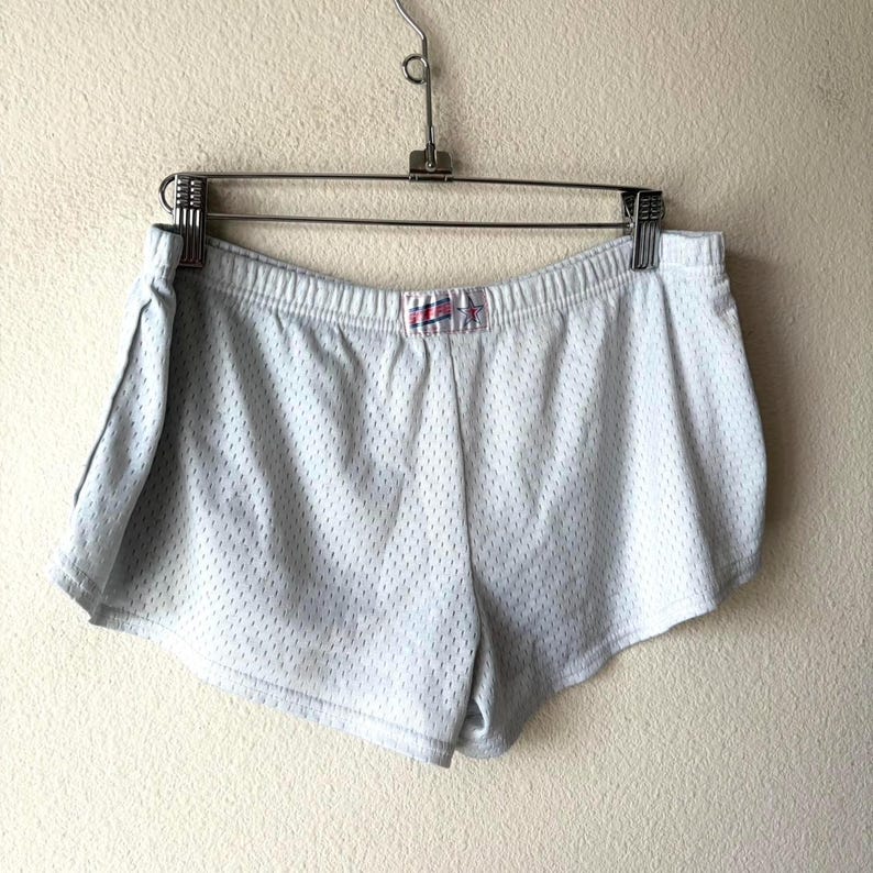 May include: White athletic shorts made of a breathable mesh fabric with an elastic waistband. A small rectangular label is centered on the front waistband featuring a blue and red striped design with a star graphic and the text "USA".