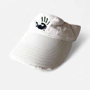 May include: White visor with a distressed brim. The visor features an embroidered logo of a handprint and the word "Nada" in dark green and black. The visor is made of a soft, textured fabric.