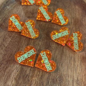 Può includere: Ciondoli in resina glitterata a forma di cuore arancione con la scritta "SHOWGIRL" in lettere verdi. I ciondoli sono sparsi su una superficie di legno. I glitter sono multicolori.