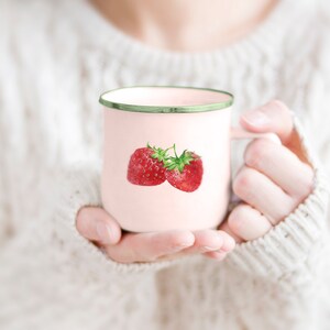 Pode incluir: Uma caneca de esmalte rosa com uma borda verde apresenta uma ilustra&ccedil;&atilde;o de aquarela de dois morangos vermelhos. A caneca est&aacute; sendo segurada por uma pessoa que est&aacute; vestindo um su&eacute;ter branco.