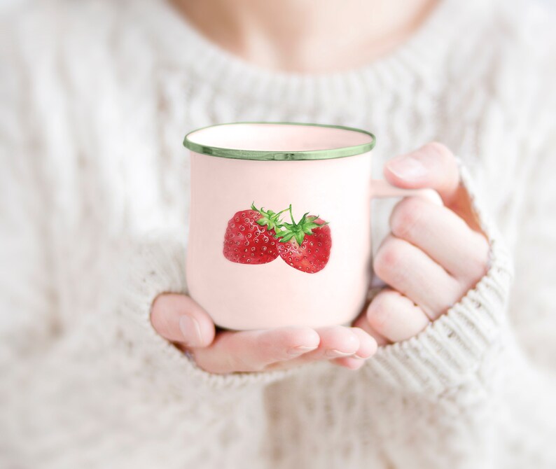 Pode incluir: Uma caneca de esmalte rosa com uma borda verde apresenta uma ilustra&ccedil;&atilde;o de aquarela de dois morangos vermelhos. A caneca est&aacute; sendo segurada por uma pessoa que est&aacute; vestindo um su&eacute;ter branco.