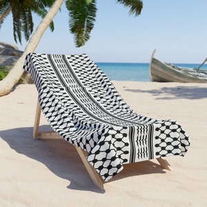 Può includere: Un telo da spiaggia a motivi bianco e nero drappeggiato su una sedia da spiaggia in legno su una spiaggia sabbiosa. Il telo ha un design tradizionale palestinese keffiyeh.