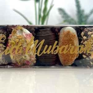 May include: A clear plastic box containing five chocolate-covered dates. The dates are decorated with nuts, white frosting, and edible flowers. The words "Eid Mubarak" are written in gold script on the box.