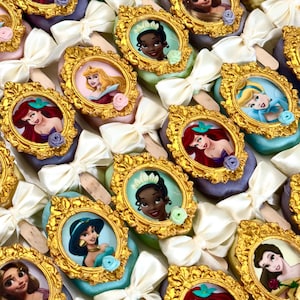 May include: An assortment of colorful, decorated cake pops featuring various Disney princess portraits in ornate gold frames. Each pop is adorned with a white bow and a small floral accent, creating a festive and elegant presentation.