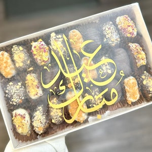 May include: A clear rectangular box filled with assorted dates, some coated in white, brown, and gold toppings. The box has a gold script design on the lid. The dates are arranged in individual brown paper cups.