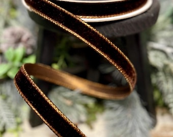 May include: A spool of dark brown velvet ribbon featuring metallic gold edges. The ribbon is wound around a black circular spool, with a length of the velvet material extending downward, showcasing the soft texture and the shimmering gold trim.