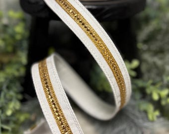 May include: White ribbon with gold beaded trim. The ribbon is spooled on a roll and also shown unraveled. The gold trim is made of small, closely spaced beads. The ribbon is on a black surface with greenery in the background.