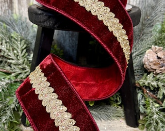 May include: A spool of deep red velvet ribbon featuring a metallic gold embroidered leaf pattern running along the center. The ribbon is wound on a spool and draped downward, showcasing the soft texture of the velvet and the intricate gold stitching.
