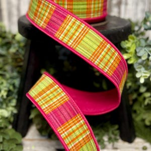 May include: A roll of decorative ribbon with a plaid pattern in shades of green, orange, and pink. The ribbon has a bright pink border and is displayed on a small black stool. The ribbon is likely used for crafts or gift wrapping.