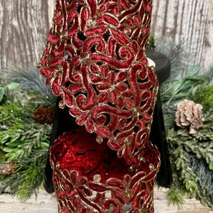 May include: A spool of wide ribbon featuring a deep red velvet texture with intricate gold metallic embroidery in a floral and scroll pattern. The ribbon has a scalloped edge and is wrapped around a central core, displaying a continuous design.