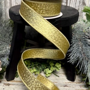 May include: A spool of gold ribbon with a textured, metallic finish. The ribbon is displayed on a small black stool, with a portion of the ribbon unrolled and draped. The ribbon is approximately 1 inch wide, ideal for crafts and gift wrapping.