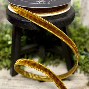 May include: A spool of brown velvet ribbon with a gold trim sits atop a small black stool. A length of the ribbon is unraveled and curves across the foreground. The ribbon is a rich, warm brown color, and the gold trim adds a touch of elegance.