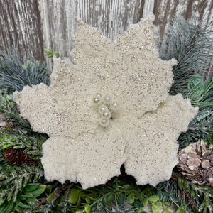 May include: A cream-colored artificial poinsettia flower with glittery petals and a cluster of small, round, pearl-like beads in the center. The flower is surrounded by green pine needles, foliage, and a small brown pinecone.