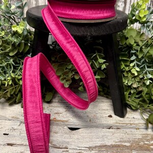 Puede incluir: Un carrete de cinta de terciopelo fucsia vibrante se encuentra sobre un peque&ntilde;o taburete negro, con un tramo de cinta que se despliega en una curva elegante. La textura afelpada y el color intenso de la cinta se destacan sobre un fondo de vegetaci&oacute;n y madera envejecida.