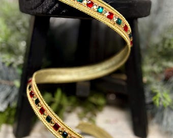 May include: A gold-colored ribbon featuring a decorative trim of alternating square red and green gems interspersed with small gold beads. The ribbon is coiled and draped, showcasing a metallic finish and a structured, woven fabric texture.