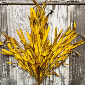 May include: A bundle of artificial yellow and brown leaves, held by a hand, against a weathered wood background. The leaves are long and slender, with a mix of bright yellow and darker brown tones, creating a natural, autumnal aesthetic. Ideal for home decor.