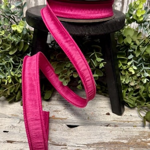 May include: A spool of vibrant fuchsia velvet ribbon sits atop a small black stool. The ribbon is unwound, creating a flowing, curved line. The background features greenery and a weathered wooden surface, adding a touch of rustic charm to the scene.